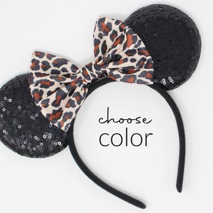 May include: A black headband with two round ears covered in black sequins and a central bow featuring a tan, brown, and black leopard print pattern. The text "choose color" is printed in black in the center of the headband.