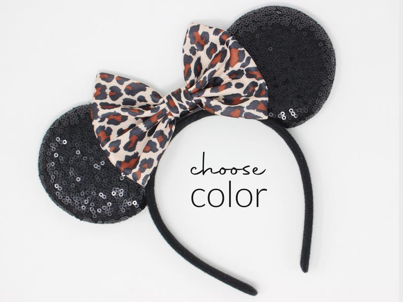 May include: A black headband with two round ears covered in black sequins and a central bow featuring a tan, brown, and black leopard print pattern. The text "choose color" is printed in black in the center of the headband.