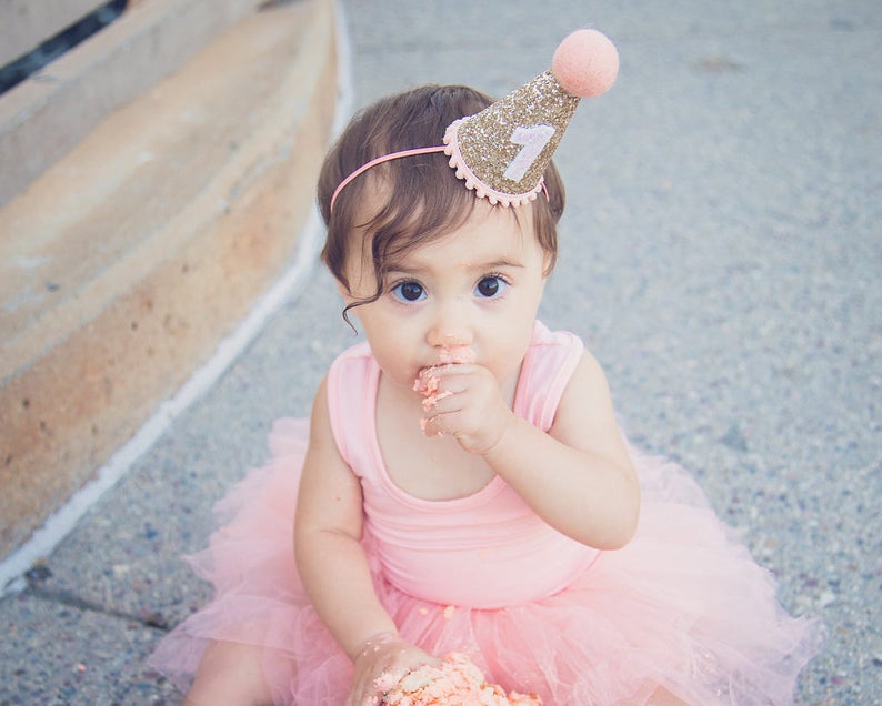 May include: A young child wearing a pink tutu and a gold glitter birthday hat with the number "1" on it. The child is eating cake and has frosting on their face.