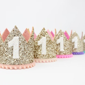 May include: A row of gold glitter crowns with white felt number "1" on each. The crowns have pink, purple, blue, and turquoise pom-pom trim.