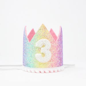 May include: A crown featuring a rainbow-colored glitter finish with a large white number 3 centered on the front. The base of the crown is trimmed with a white pom-pom fringe, and it includes thin white elastic straps on the sides for securing.