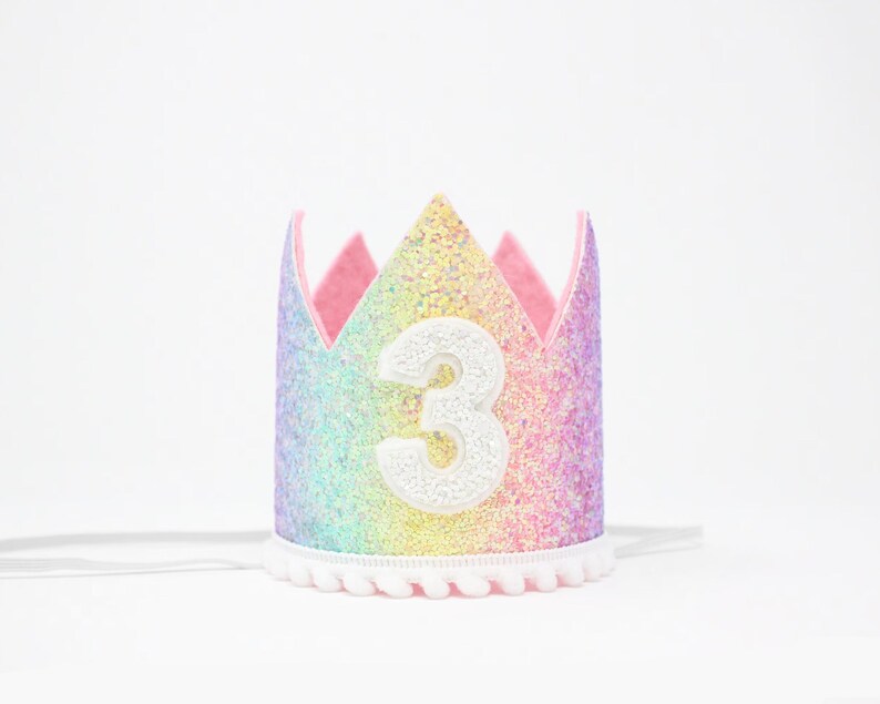 May include: A crown featuring a rainbow-colored glitter finish with a large white number 3 centered on the front. The base of the crown is trimmed with a white pom-pom fringe, and it includes thin white elastic straps on the sides for securing.