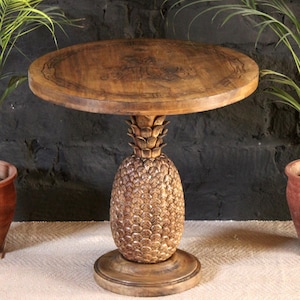 Puede incluir: Una mesa redonda de madera con una base de piña tallada. La tapa de la mesa tiene un diseño decorativo y es de color marrón.