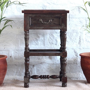 Może przedstawiać: Brązowy drewniany stolik nocny z jedną szufladą i dolną półką. Stolik ma toczone nogi i ozdobny detal na dolnej półce.
