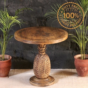 May include: A round wooden side table featuring a pedestal base carved in the shape of a pineapple with a textured surface. The tabletop has a smooth, circular grain pattern. The table is positioned between two potted green plants.