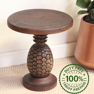 May include: A dark brown wooden side table featuring a circular top and a base carved in the shape of a pineapple. The table has a textured, natural wood grain finish throughout. A green circular stamp with the text DUTY FREE 100% TARIFF PREPAID.