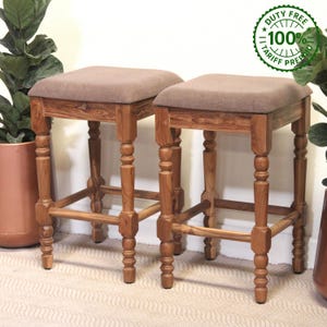 May include: Two wooden bar stools with square, light brown upholstered seats. Each stool features four turned legs with a natural wood grain finish and horizontal support bars connecting the legs for stability and structural support.