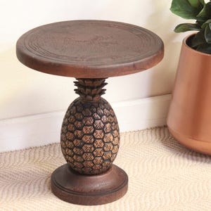 Puede incluir: Una mesa auxiliar de madera marrón oscuro con una tapa redonda y una base en forma de piña. La mesa tiene un diseño tallado en la parte superior y la base de piña. La mesa mide aproximadamente 51 cm de altura.