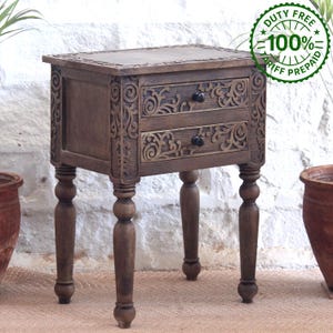 May include: A brown wooden bedside table featuring two drawers with intricate carved scrollwork patterns on the drawer fronts and side panels. The piece stands on four turned legs and includes small black knobs on each drawer for opening.