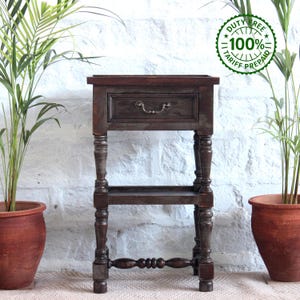May include: A dark brown wooden side table featuring a single drawer with a curved metal handle, a lower open shelf, and four turned legs. The table has a rectangular top and a sturdy frame construction with visible wood grain throughout the piece.