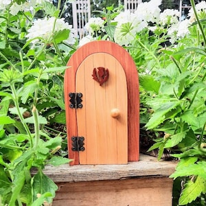 May include: A small wooden fairy door with a brown door and a red wooden heart design. The door is surrounded by white flowers and green leaves.