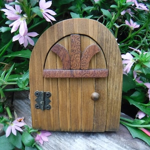 May include: A small wooden door with a rounded top and a brown finish. The door has a simple design with a decorative arch and a small knob. The door is surrounded by green foliage and pink flowers.
