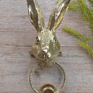 May include: A gold-toned rabbit head door knocker with a rope-style ring. The knocker is mounted on a wooden surface.