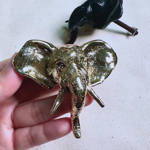 Peut inclure: Deux boutons de meuble en forme d'éléphant. L'un est doré, avec des détails, dont une trompe et de grandes oreilles. L'autre est noir et possède une vis pour l'installation. Les boutons sont sur une surface claire.