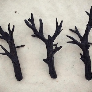 Puede incluir: Tres ganchos de pared de metal negro con forma de ramas de árbol estilizadas con un solo tornillo en cada uno.