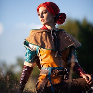 Peut inclure: Une femme aux cheveux roux portant un gilet en cuir marron avec une ceinture bleue, une chemise bleu sarcelle et marron et un pantalon marron. Elle est assise dans un champ d'herbe avec un ciel bleu en arrière-plan.