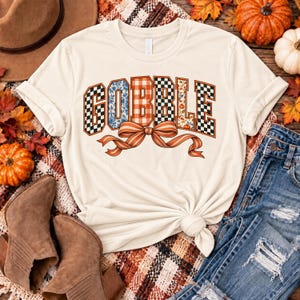 Könnte beinhalten: Ein cremefarbenes Kurzarm-T-Shirt mit dem Wort GOBBLE in großen Blockbuchstaben. Jeder Buchstabe hat ein einzigartiges Muster, darunter blaue Blumen, orangefarbene Karos, ein schwarz-weißes Schachbrettmuster und herbstliche Blätter.
