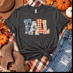 Könnte beinhalten: Ein dunkelgraues Kurzarm-T-Shirt mit dem Wort FALL in großen Blockbuchstaben. Jeder Buchstabe hat ein eigenes Muster: blaues Blumenmuster, cremefarbenes Blumenmuster, orange-weißes Vichy-Karo und ein cremefarbenes Karomuster mit kleinen orangefarbenen Blumen.