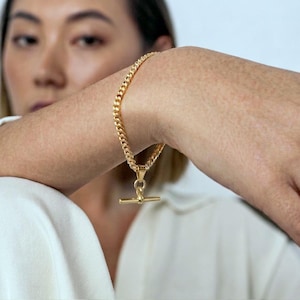 Peut inclure: Un bracelet chaîne doré avec un fermoir en T. Le bracelet présente un design de chaîne épais et texturé. Le fermoir est une simple barre qui coulisse dans une boucle. Le bijou est présenté sur le bras d'une personne.