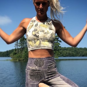 Pode incluir: Uma mulher vestindo um top branco curto com estampa de folhas amarelas e verdes e calças de ioga cinza com estampa de folhas pretas. Ela está em pé em frente a um lago.