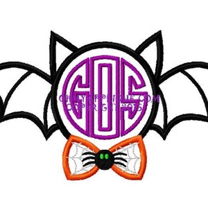 May include: A purple monogram in the shape of a bat with black wings and a black and orange bow tie with a spider in the center.