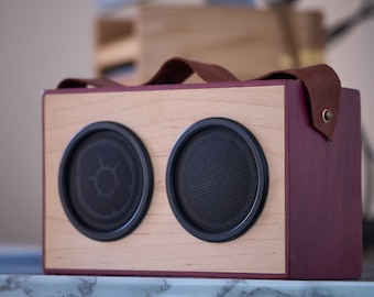 May include: A wooden speaker with two round speakers and a brown leather strap. The speaker is made of light wood with a dark red finish on the side.
