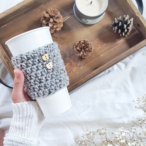 May include: A gray crocheted coffee cup cozy with two wooden buttons. The cozy is on a white paper cup. The cup is on a wooden tray with pine cones and a candle.
