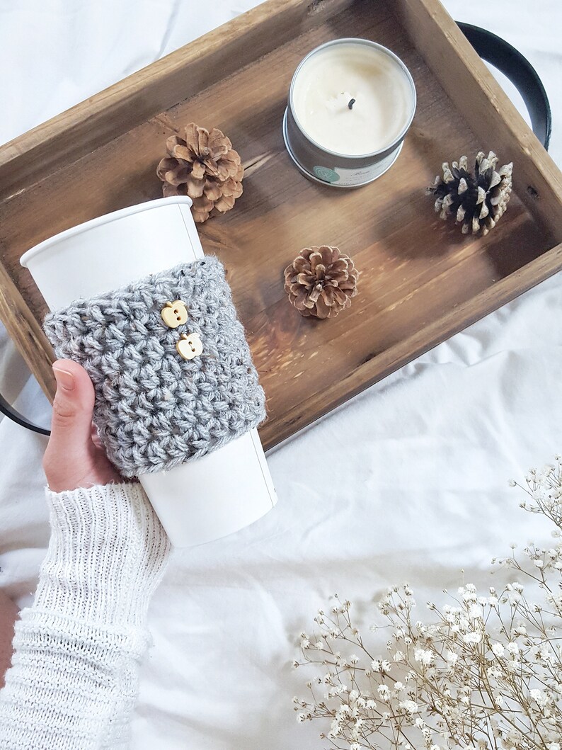 May include: A gray crocheted coffee cup cozy with two wooden buttons. The cozy is on a white paper cup. The cup is on a wooden tray with pine cones and a candle.