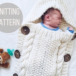 May include: Knitting pattern for a white hooded baby wrap with wooden buttons. The wrap is made with a cable knit stitch and has a hood that can be pulled over the baby's head. The wrap is shown being worn by a baby.
