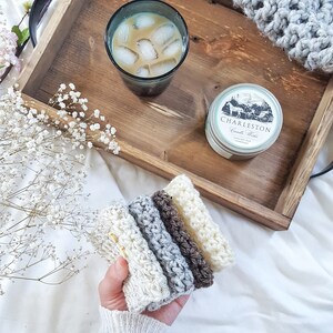 May include: A set of five crocheted dishcloths in various shades of brown, gray, and white. The dishcloths are stacked on top of each other and are being held by a hand. The dishcloths are on a white surface.
