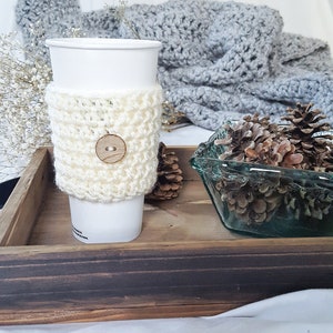 May include: A white crocheted coffee cup cozy with a wooden button. The cozy is on a wooden tray with a glass bowl of pine cones.