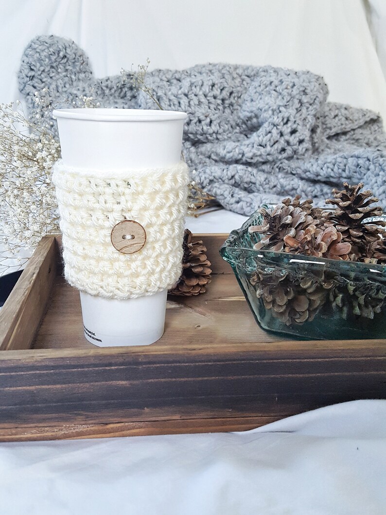 May include: A white crocheted coffee cup cozy with a wooden button. The cozy is on a wooden tray with a glass bowl of pine cones.