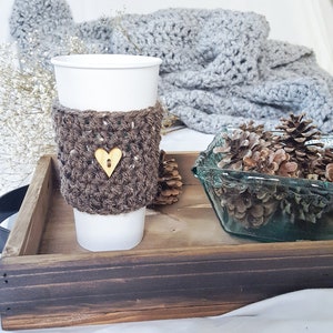 May include: A brown crocheted coffee cup cozy with a wooden heart button. The cozy is on a white paper cup and is sitting on a wooden tray with pine cones in a glass bowl.