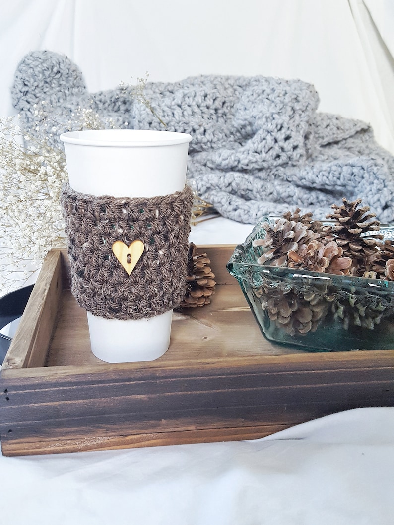 May include: A brown crocheted coffee cup cozy with a wooden heart button. The cozy is on a white paper cup and is sitting on a wooden tray with pine cones in a glass bowl.