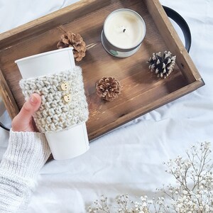 May include: A white coffee cup with a crocheted beige cozy with two wooden buttons. The cup is in a wooden tray with a white candle, pine cones, and a small bouquet of white baby's breath.