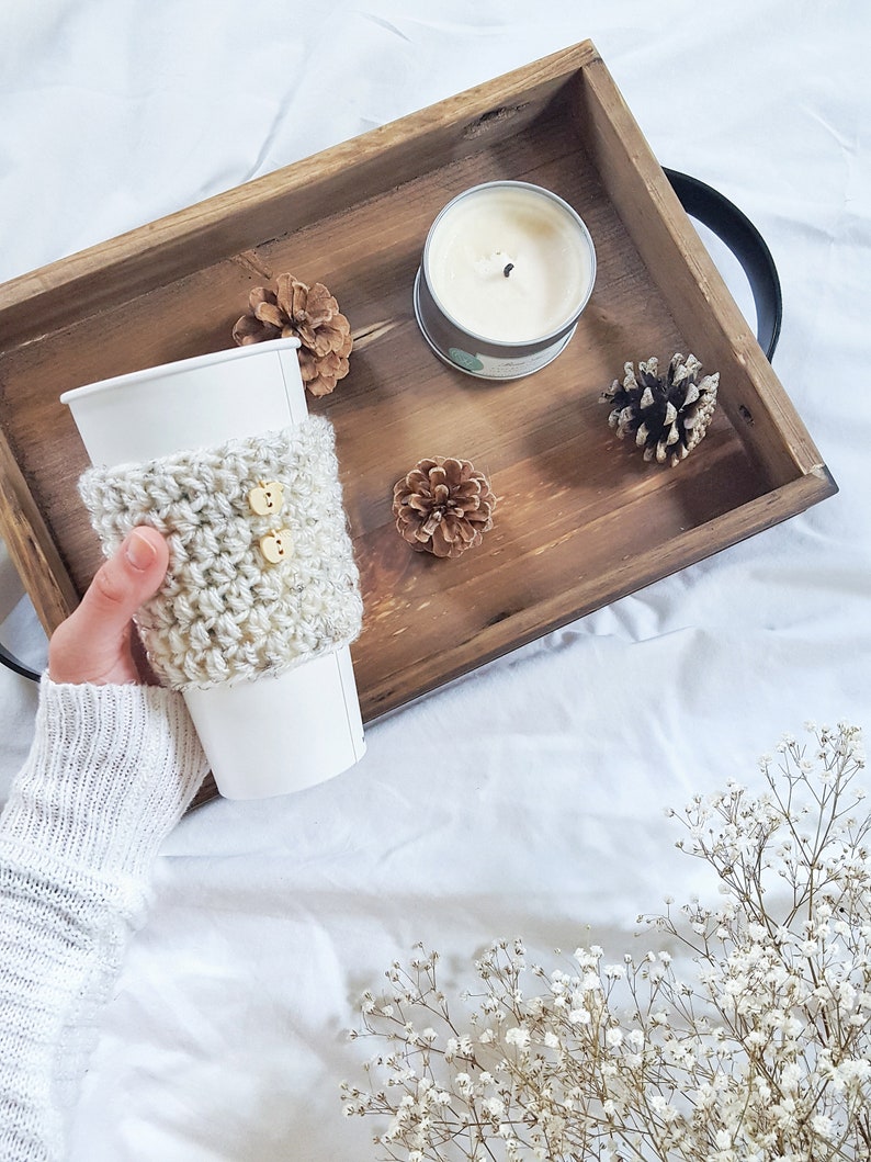 May include: A white coffee cup with a crocheted beige cozy with two wooden buttons. The cup is in a wooden tray with a white candle, pine cones, and a small bouquet of white baby's breath.