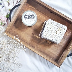 May include: A wooden tray with a crocheted square and a round tin with the text "Charleston Candle Works" on it.