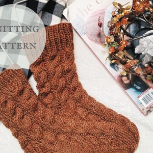 May include: A pair of brown knitted socks with a cable knit pattern. The socks are lying on a white fluffy surface with a black and white checked blanket in the background.
