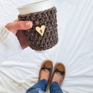 May include: A brown crocheted coffee cup cozy with a wooden heart button. The cozy is on a white paper cup. The person is wearing blue jeans and brown fuzzy slippers.