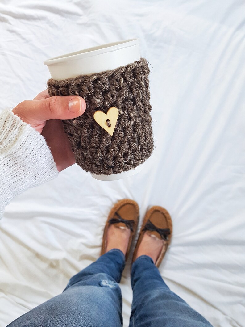 May include: A brown crocheted coffee cup cozy with a wooden heart button. The cozy is on a white paper cup. The person is wearing blue jeans and brown fuzzy slippers.
