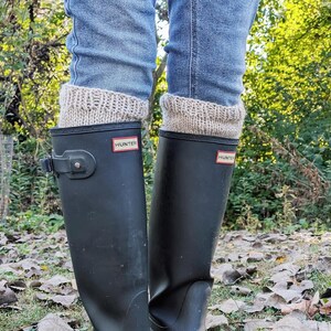 Könnte beinhalten: Schwarze Gummiregenstiefel mit Hunter-Logo und beige gestrickten Stiefelmanschetten. Die Stiefel werden mit blauen Jeans getragen. Der Hintergrund zeigt Herbstlaub und grünes Laub.