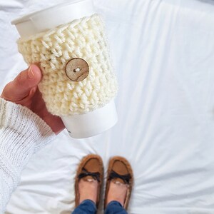 May include: A white crocheted coffee cup cozy with a wooden button. The cozy is on a white paper cup. The cup is being held by a person wearing blue jeans and brown fuzzy slippers.