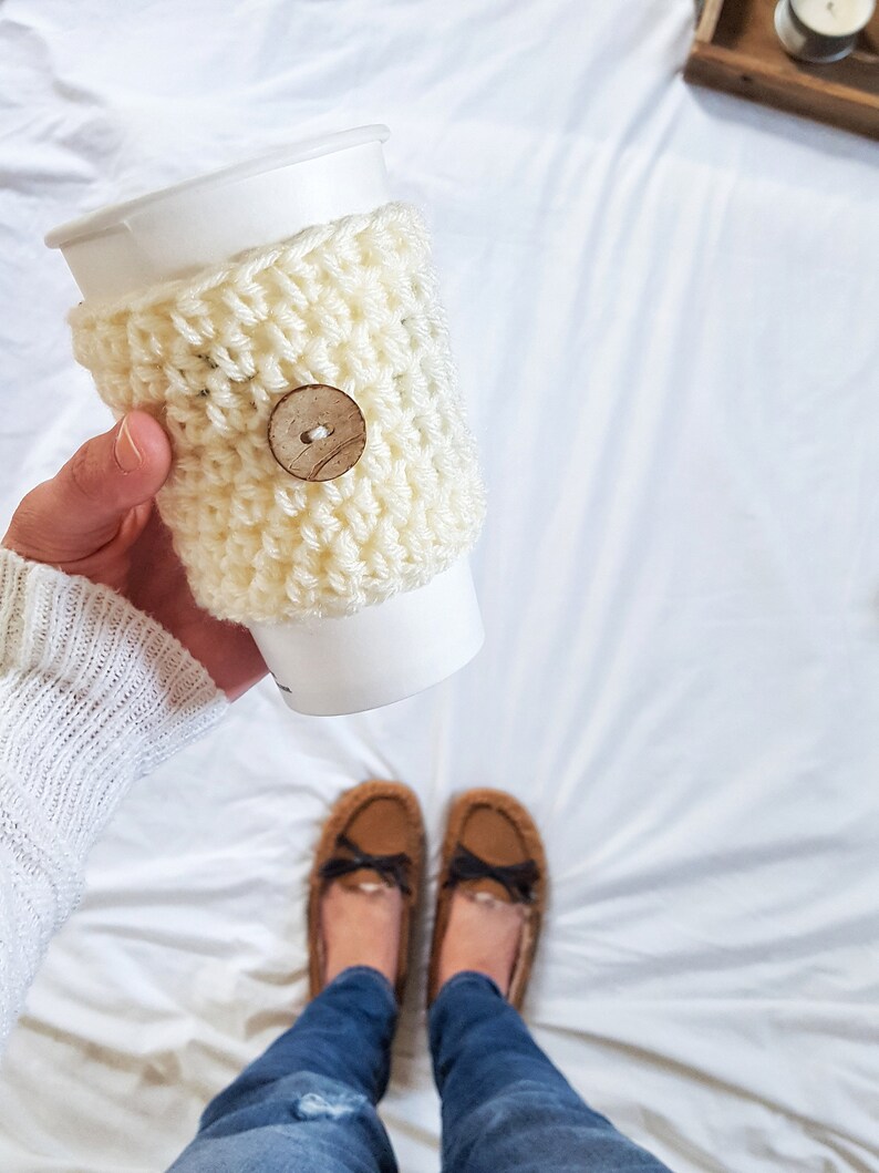 May include: A white crocheted coffee cup cozy with a wooden button. The cozy is on a white paper cup. The cup is being held by a person wearing blue jeans and brown fuzzy slippers.