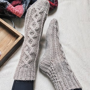 Könnte beinhalten: Ein Paar graue Stricksocken mit einem Zopfmuster. Die Socken liegen auf einer weißen, flauschigen Oberfläche.