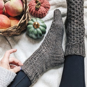 May include: A pair of brown knitted socks with a textured pattern. The socks are worn on a person's feet and legs. The person is wearing black pants. The socks are on a white fuzzy surface with a basket of apples and gourds.