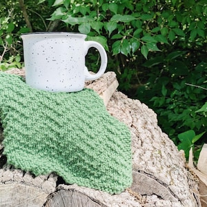 Op de afbeelding: Een witte geëmailleerde mok met zwarte spikkels en een zwarte rand staat op een groene gebreide washand. De washand heeft een textuurpatroon. De mok staat op een stapel houtblokken, met groen gebladerte op de achtergrond.
