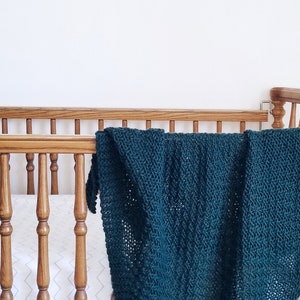May include: A dark green knitted baby blanket draped over a wooden cot with white bedding.