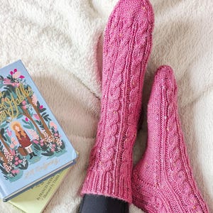 May include: A close-up of pink cable-knit socks and a book titled "Anne of Green Gables" on a white blanket. The book has a light blue cover with an illustration of a girl in a forest. The socks are a vibrant pink colour.