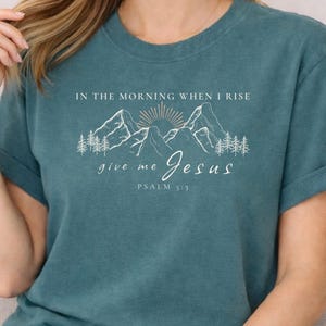 Könnte beinhalten: Türkisfarbenes T-Shirt mit weißem Text: "IN THE MORNING WHEN I RISE give me Jesus PSALM 5:3". Das Design zeigt eine Bergkette, Sonne und Bäume. Die Ärmel sind hochgekrempelt.