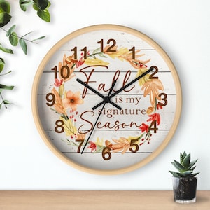 Puede incluir: Un reloj de pared redondo con marco de madera clara. La esfera tiene un fondo blanco con una corona de acuarela de hojas de otoño y el texto "Fall is my signature Season."